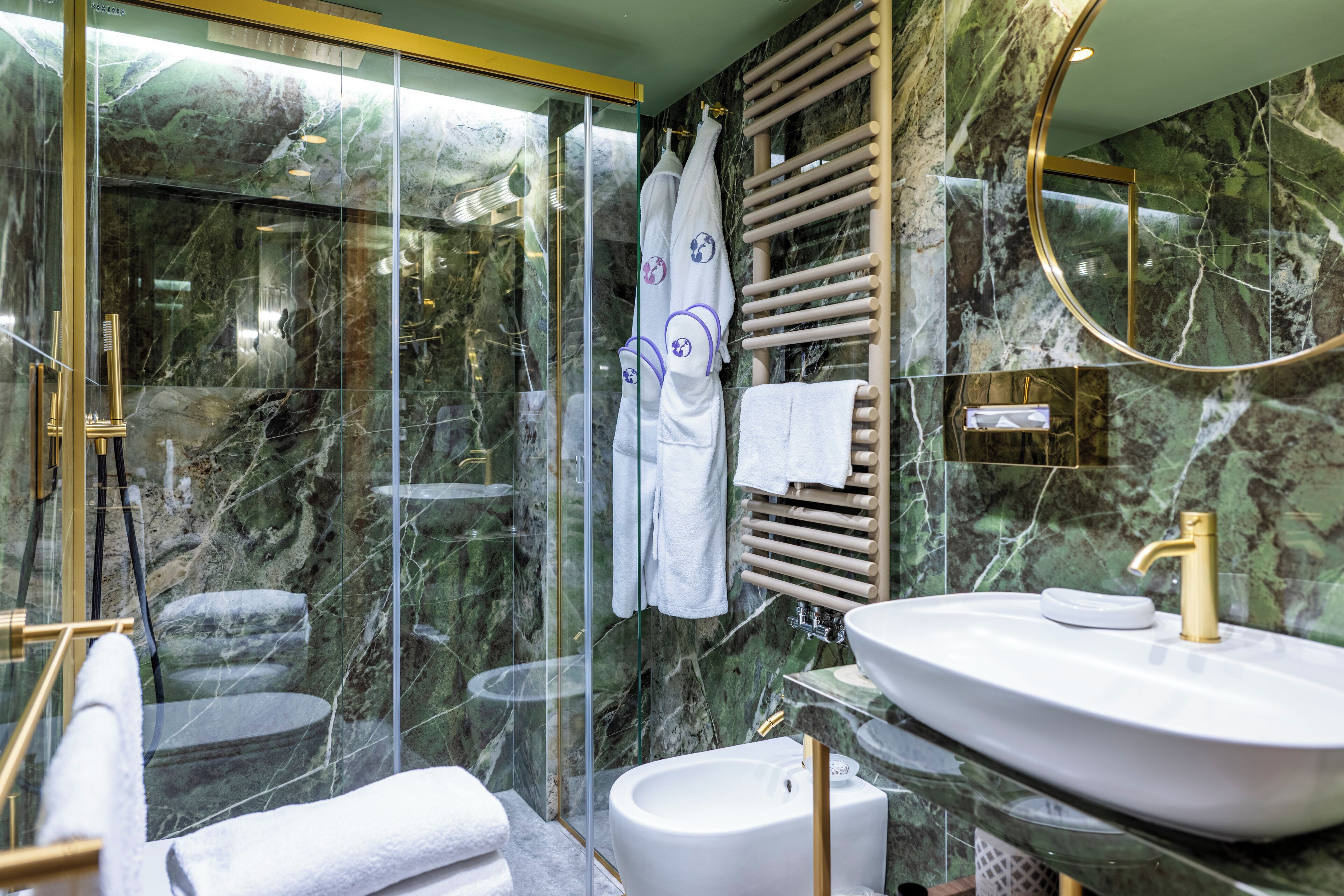A modern bathroom with a glass-enclosed shower, a marble wall, a white sink with a gold faucet, and a towel rack.
