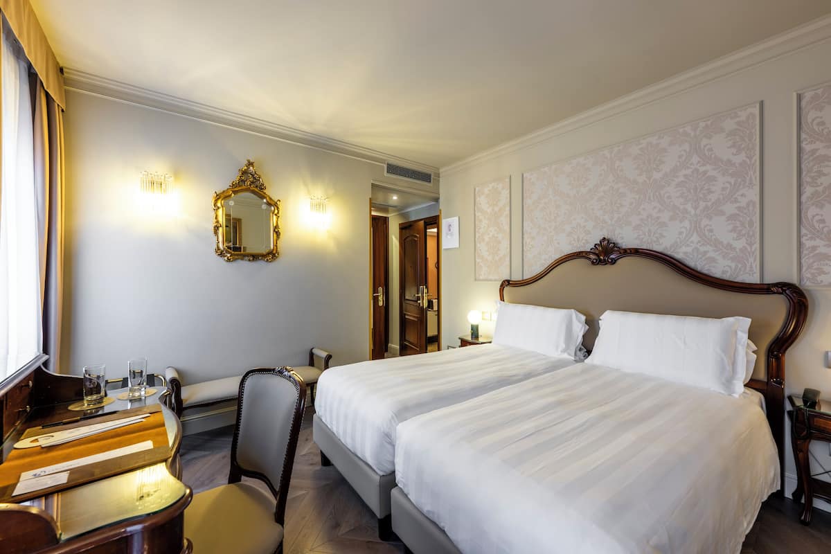A hotel room with two beds, a desk, a chair, a mirror, and a wall with patterned wallpaper.