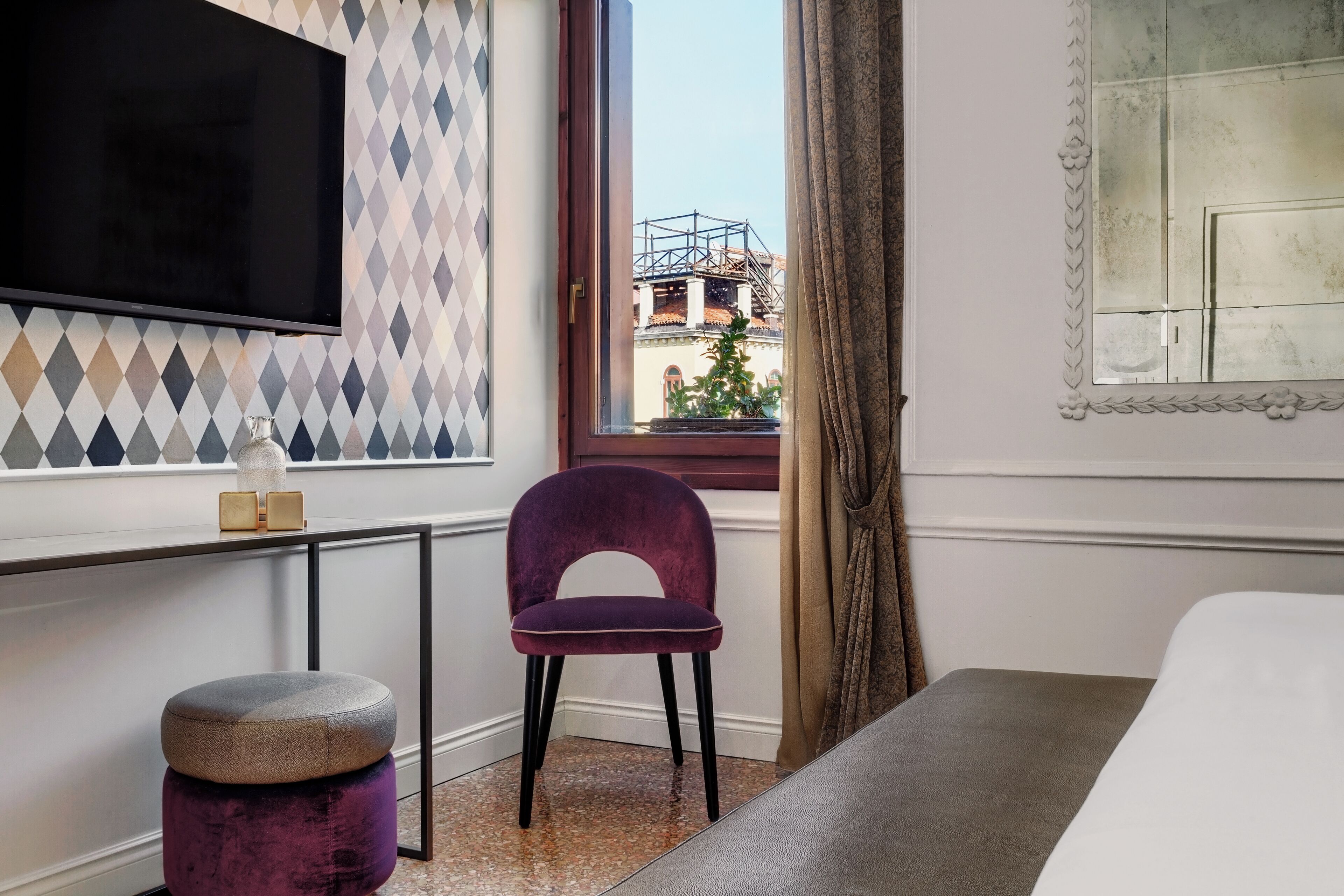 Une chambre d’hôtel avec un lit, un téléviseur fixé au mur, une fenêtre donnant sur des immeubles, une chaise et une petite table avec une lampe.