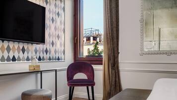 Une chambre d’hôtel avec un lit, un téléviseur fixé au mur, une fenêtre donnant sur des immeubles, une chaise et une petite table avec une lampe.
