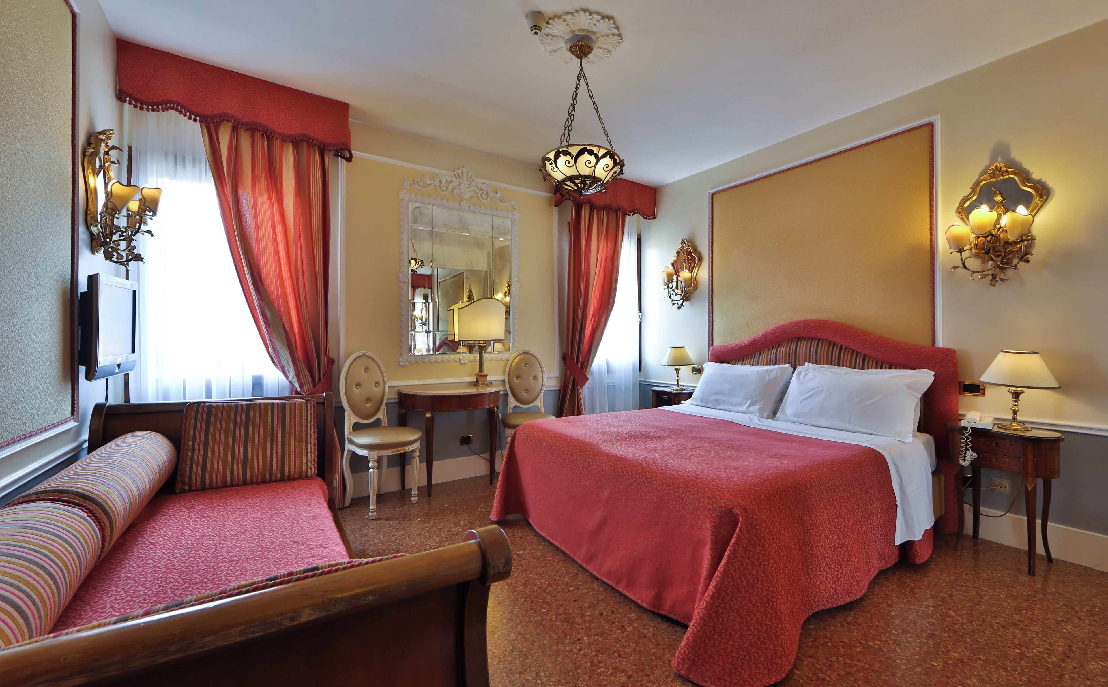 Hotelkamer met een groot bed, een zithoek met een bank en fauteuil, een bureau met stoel, en een spiegel.