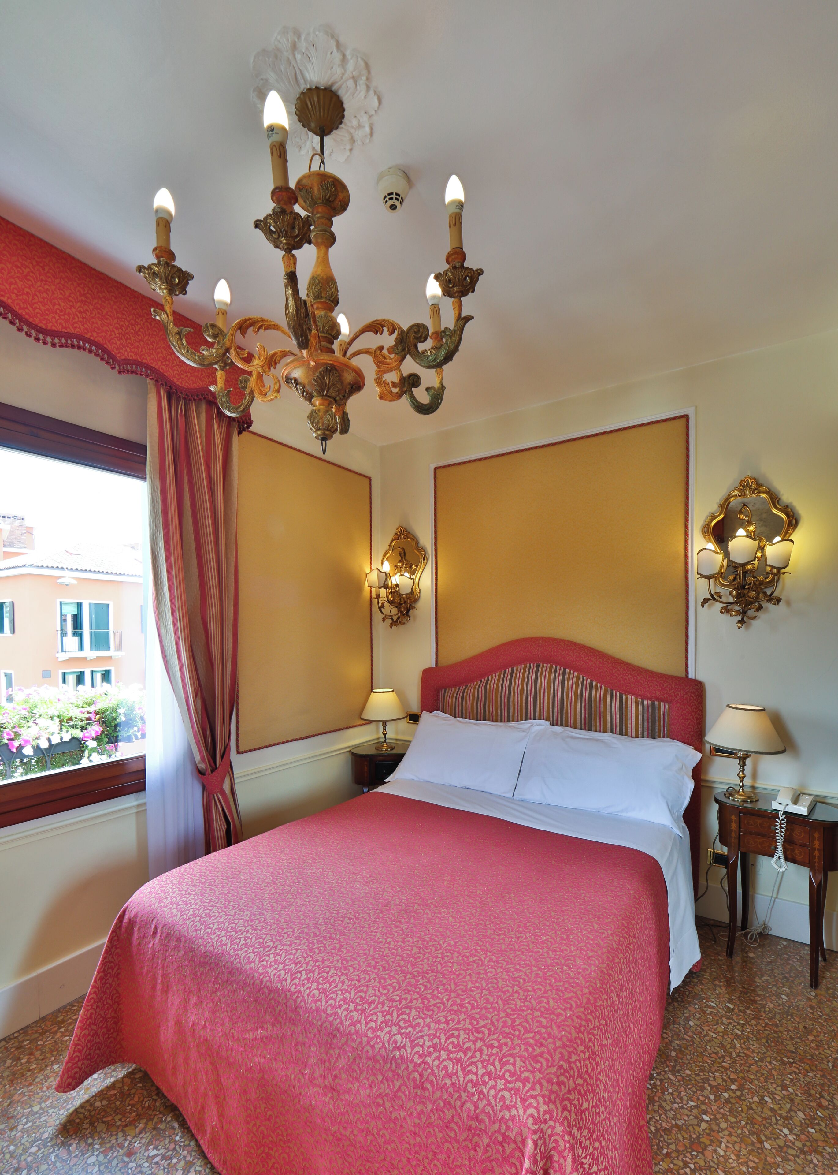 A hotel room with a large bed, a chandelier, two wall sconces, a window with curtains, and a view of buildings outside.