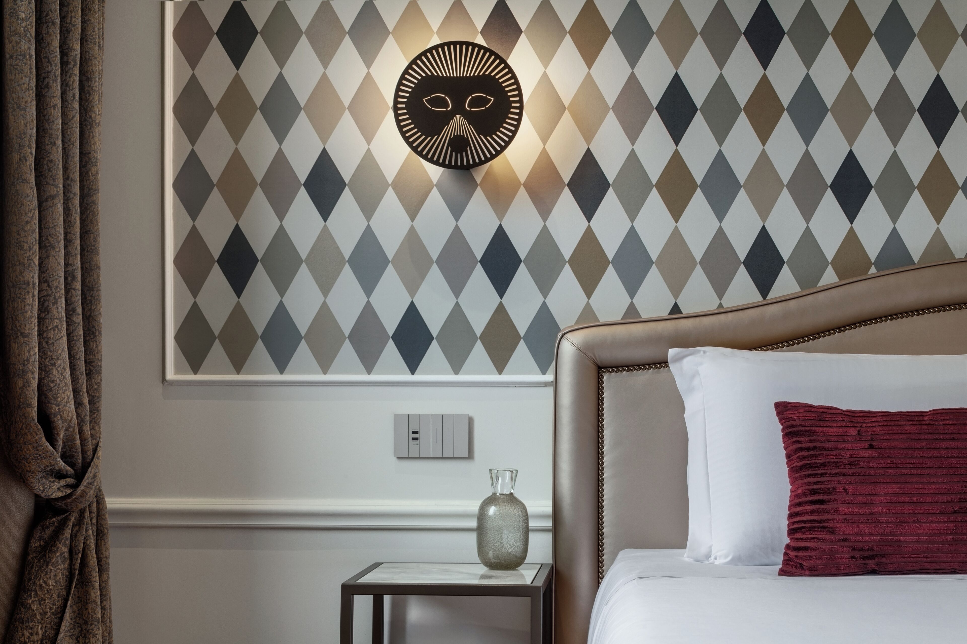 A hotel room with a patterned wall, a bed with white and red pillows, a nightstand with a vase, and a wall-mounted light fixture.