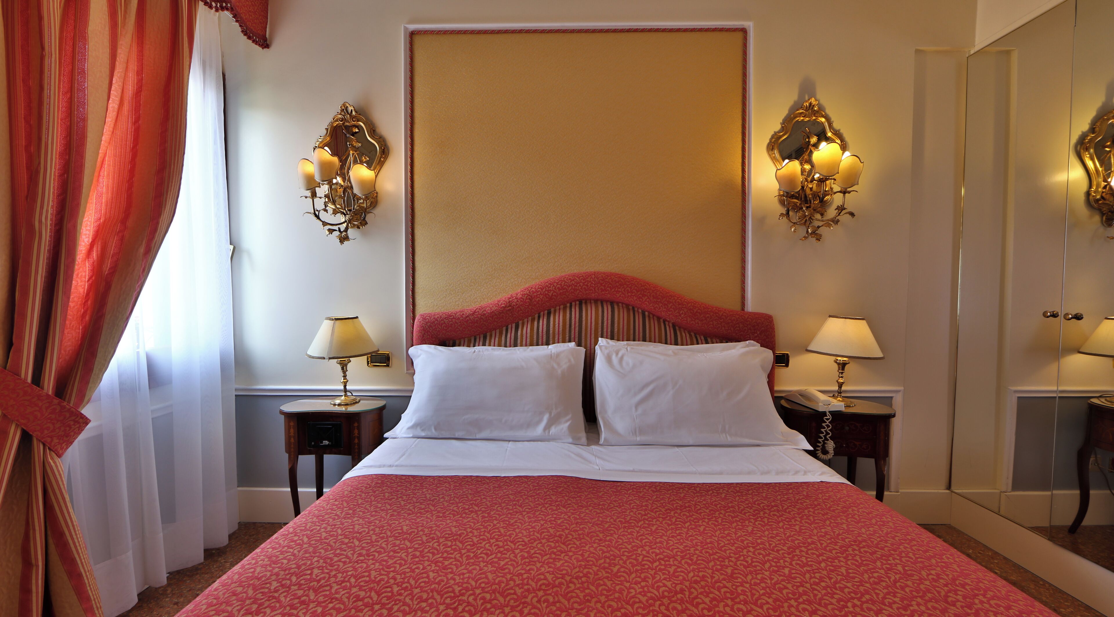 A hotel room with a large bed, two bedside tables with lamps, a large mirror, and a red curtain.