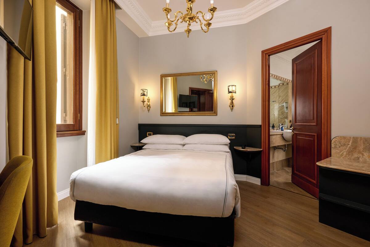 A hotel room with a large bed, a bedside table, a mirror, a chandelier, and a door leading to a bathroom.