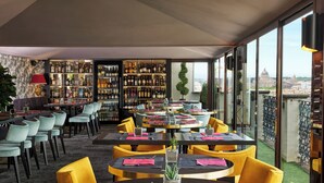 A restaurant with a bar area, tables with yellow chairs, and a view of a cityscape through large windows. - Hotel Barberini (Rome)