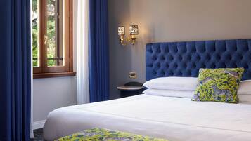Una camera d'albergo con un letto grande, una finestra con tende blu, un lampadario a parete e una testiera imbottita.