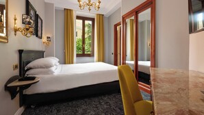 A hotel room with a bed, a desk, a chair, a chandelier, and a window with curtains. - Hotel Barberini (Rome)