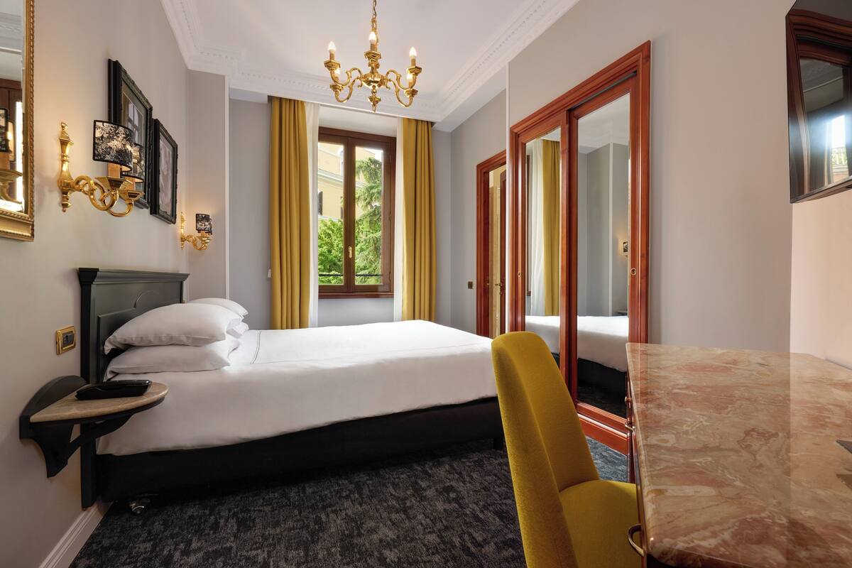 A hotel room with a bed, a desk, a chair, a chandelier, and a window with curtains.