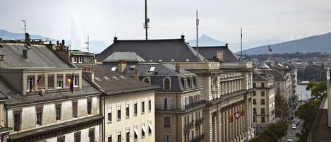 A row of historic buildings with balconies, a river in the background, and a mountain range.
