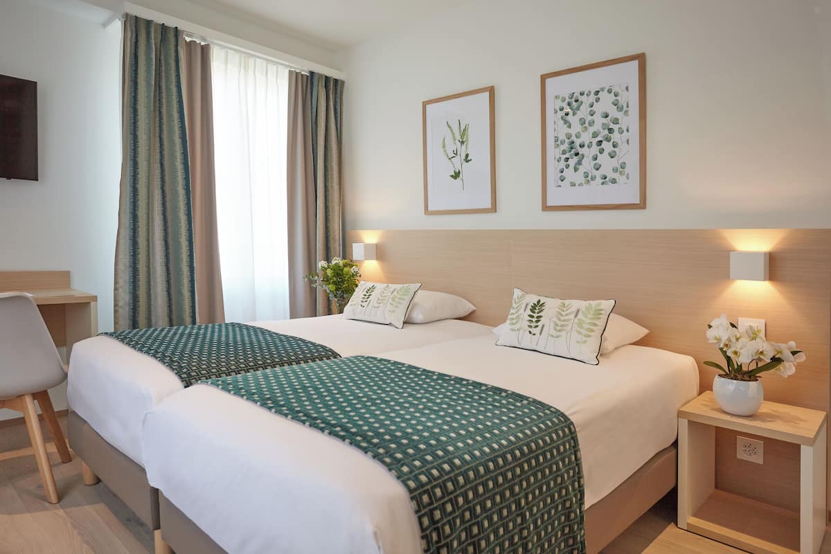 A hotel room with two beds, a desk, a chair, a nightstand with a vase of flowers, and two framed botanical prints on the wall.