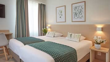 Habitación de hotel con dos camas, un escritorio, una silla, una mesita de noche con un jarrón de flores e dos láminas botánicas enmarcadas en la pared.