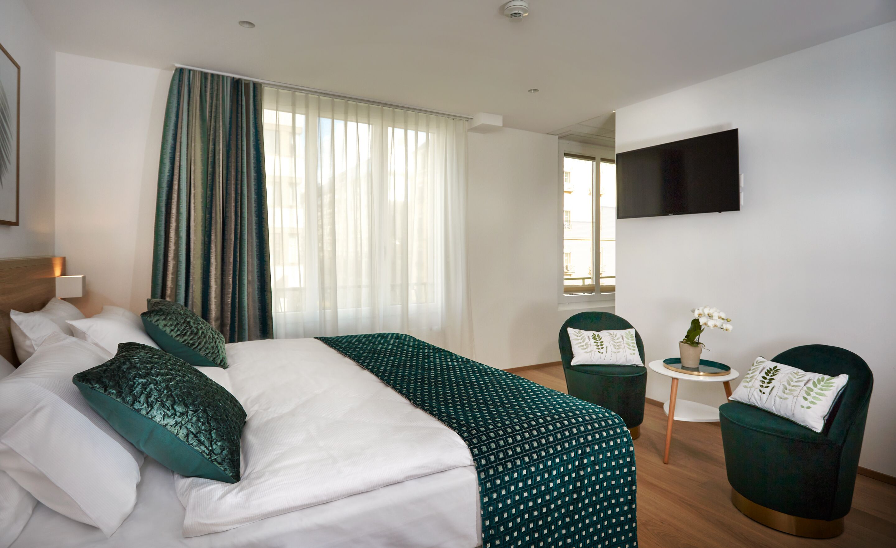 Habitación de hotel con cama, dos sillas, una mesita, televisor y ventana con cortinas.