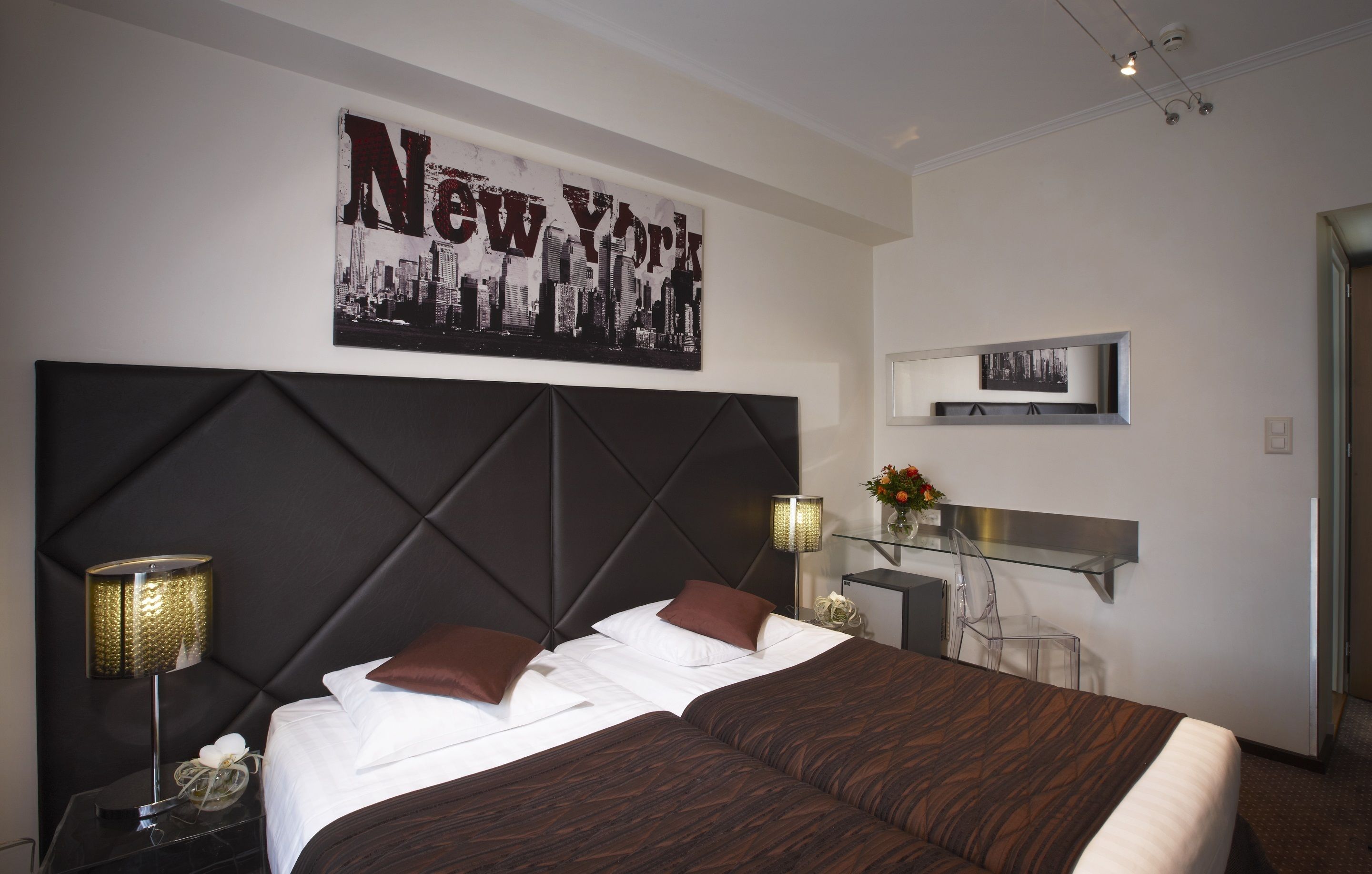 Une chambre d’hôtel avec un grand lit, une tête de lit, une œuvre d’art représentant New York accrochée au mur, une table de chevet avec une lampe et un petit bureau agrémenté d’un vase de fleurs.