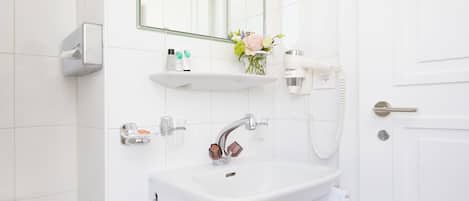 A bathroom with a white sink, a mirror, a towel rack, a hairdryer, and a shelf with toiletries.