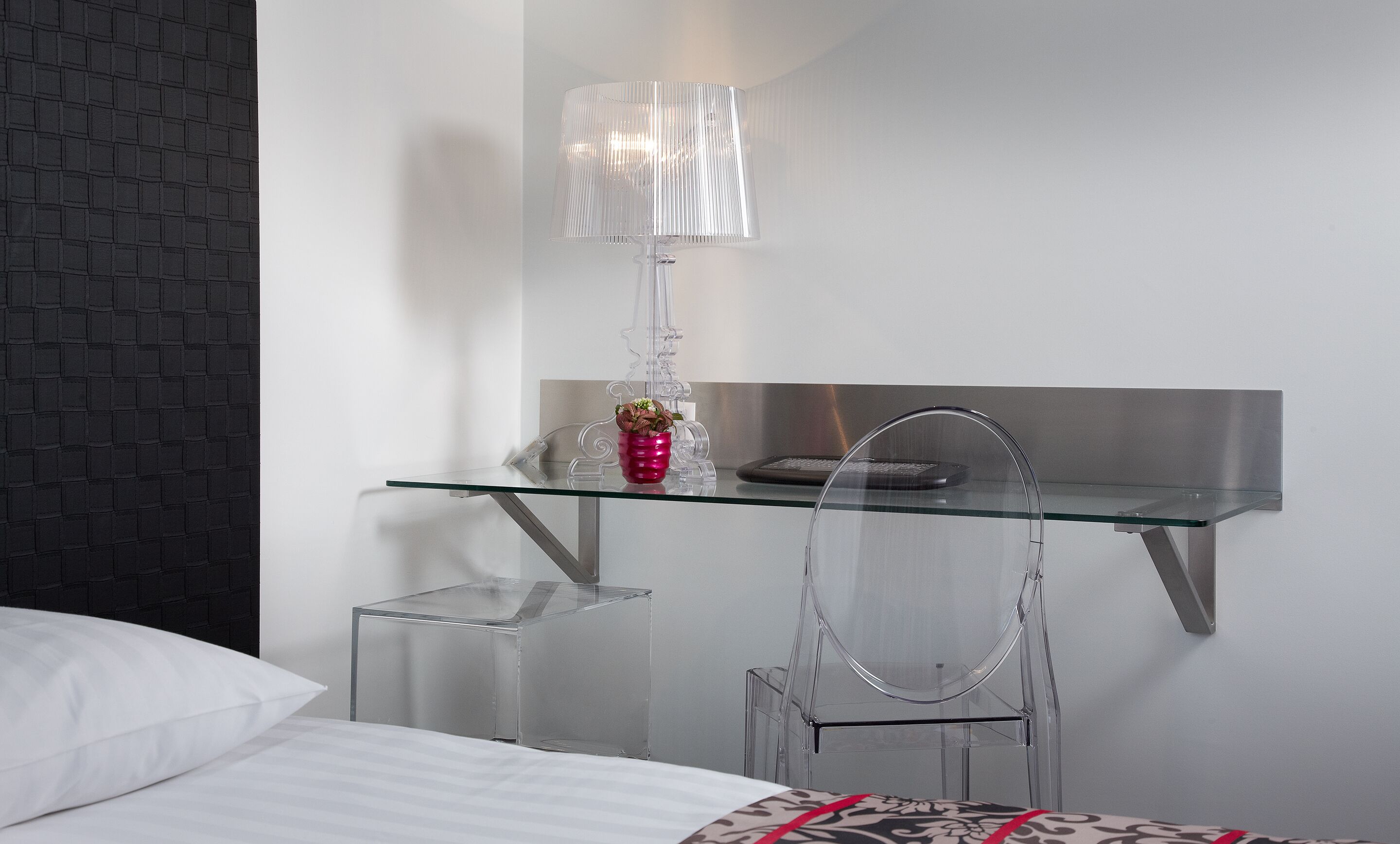 A modern hotel room with a glass desk, a clear chair, a lamp, and a bed with white bedding.