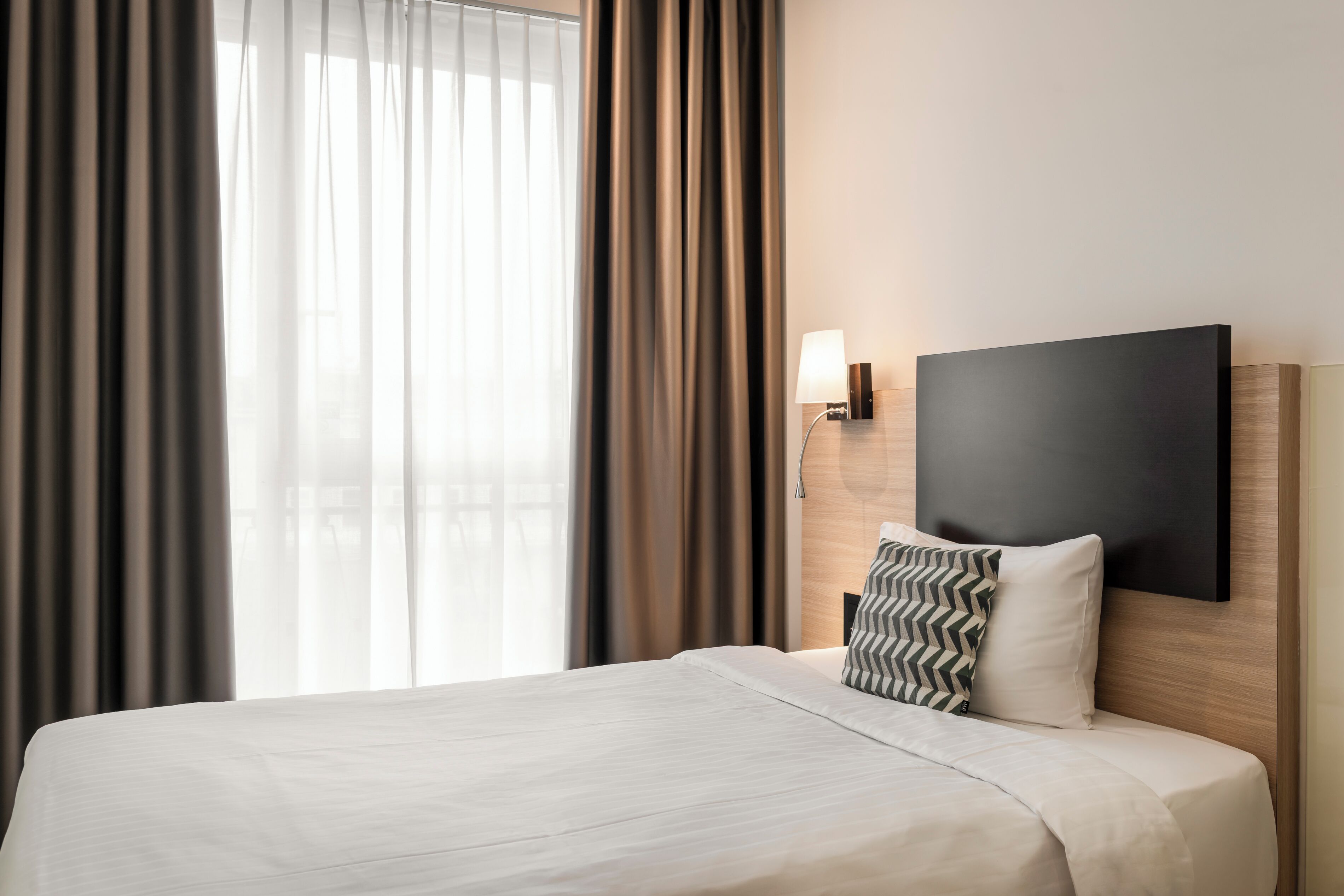 A hotel room with a bed, curtains, a wall-mounted lamp, and a painting.