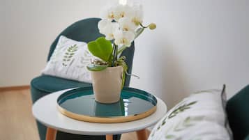 Una pequeña mesa redonda con una orquídea en maceta, una silla verde con un cojín blanco y un cojín blanco con estampado de hojas.