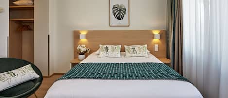 A hotel room with a bed, a chair, a nightstand, a wall-mounted picture, and a window with curtains.