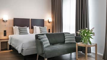 Quarto de hotel com uma cama, um sofá, uma mesa de cabeceira com um vaso de flores e uma janela com cortinas.