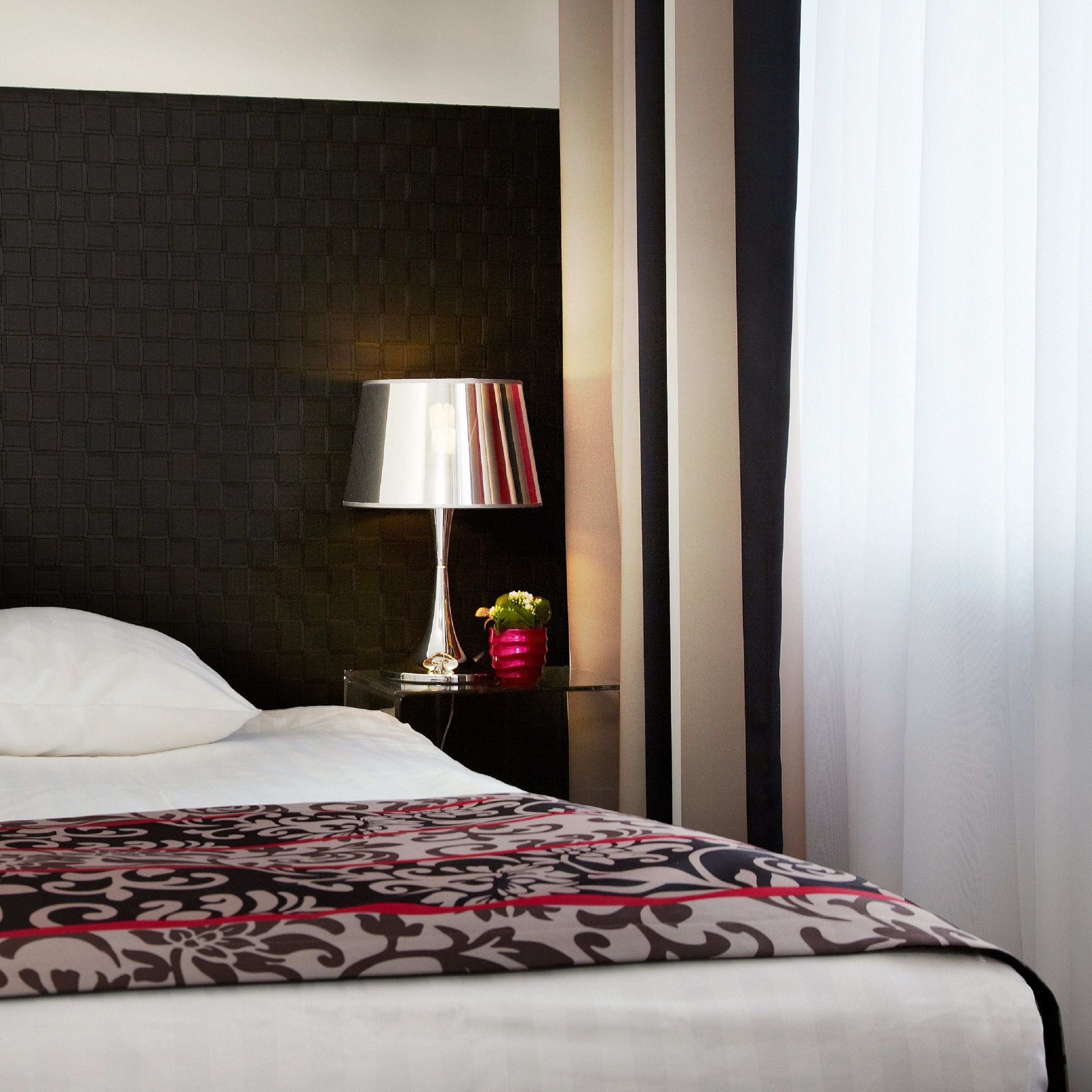 A hotel room with a bed, a nightstand with a lamp, a vase with flowers, and a dark textured wall.