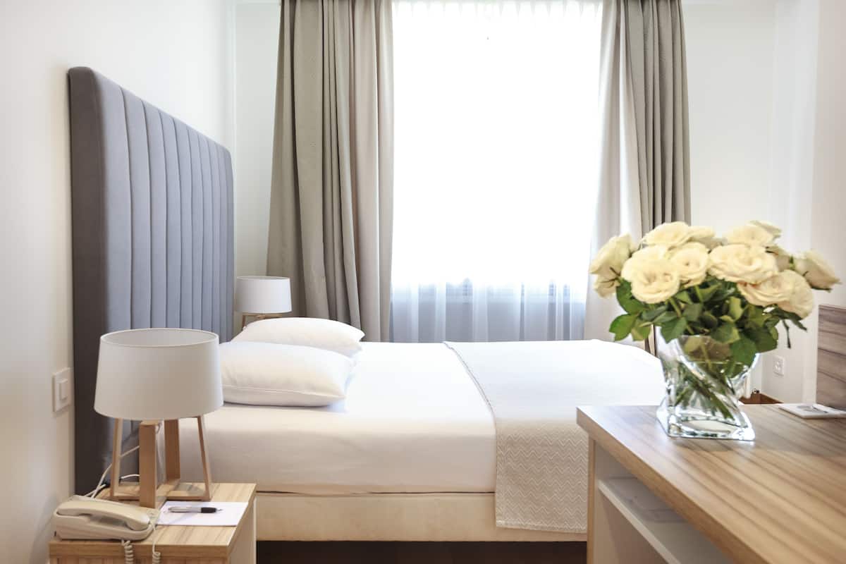A neatly made bed with a white comforter and pillows, a bedside lamp, a telephone, and a vase with white flowers on a wooden nightstand.