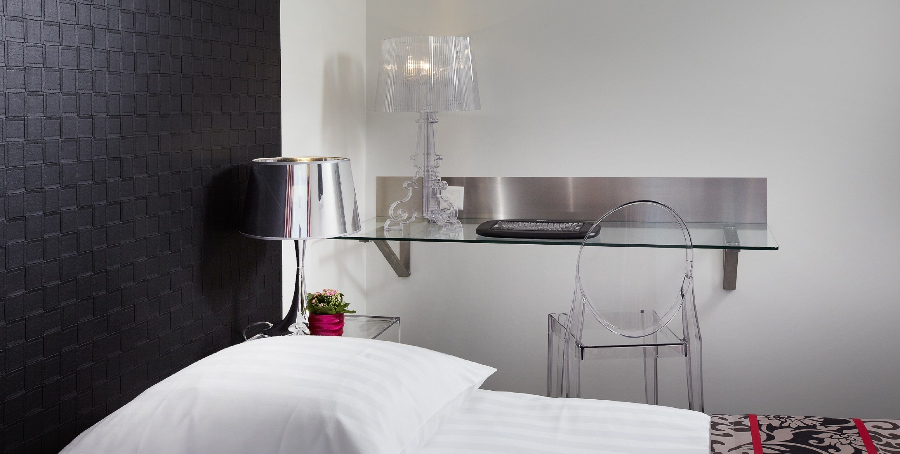 Una habitación de hotel moderna con una pared de ladrillo negro, un escritorio de vidrio con una computadora, una silla transparente y una cama blanca.