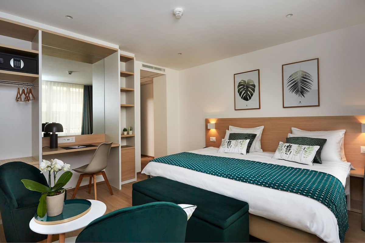 A hotel room with a bed, a desk, a chair, a small table with a plant, and a closet.