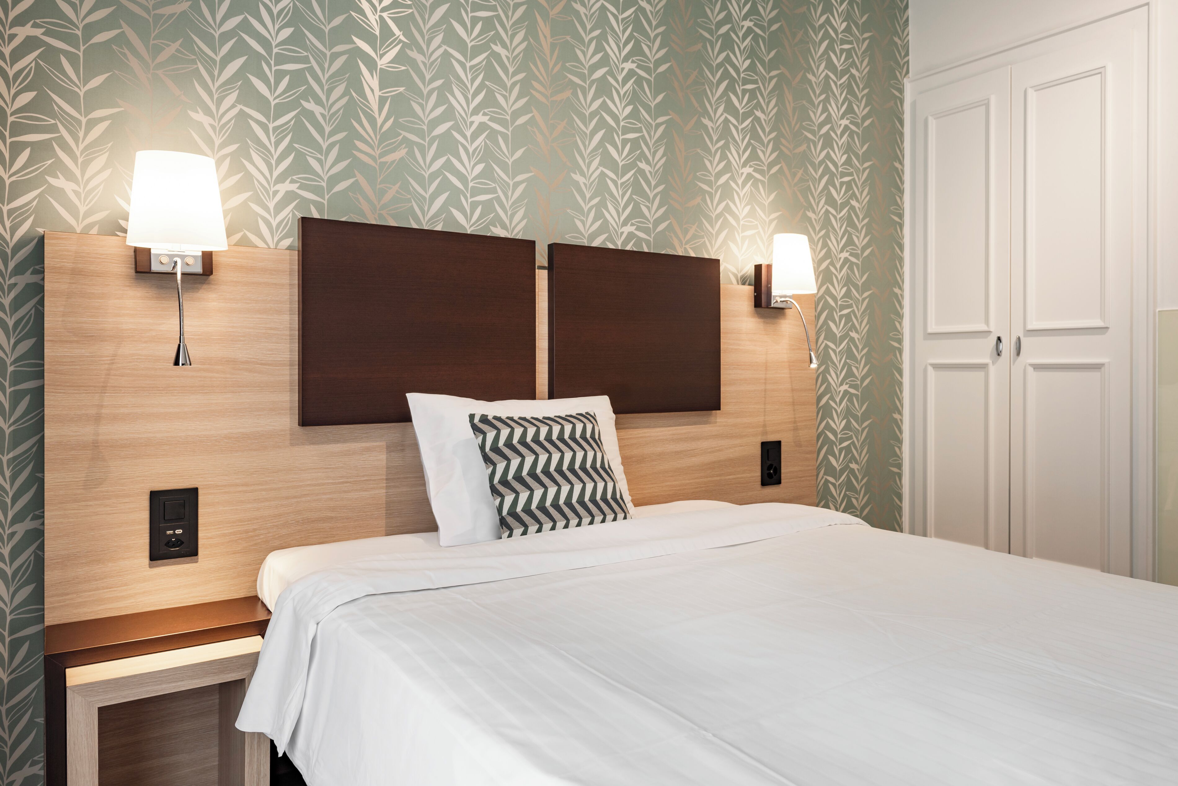 A neatly made bed with a wooden headboard, two bedside lamps, and a patterned wallpaper.