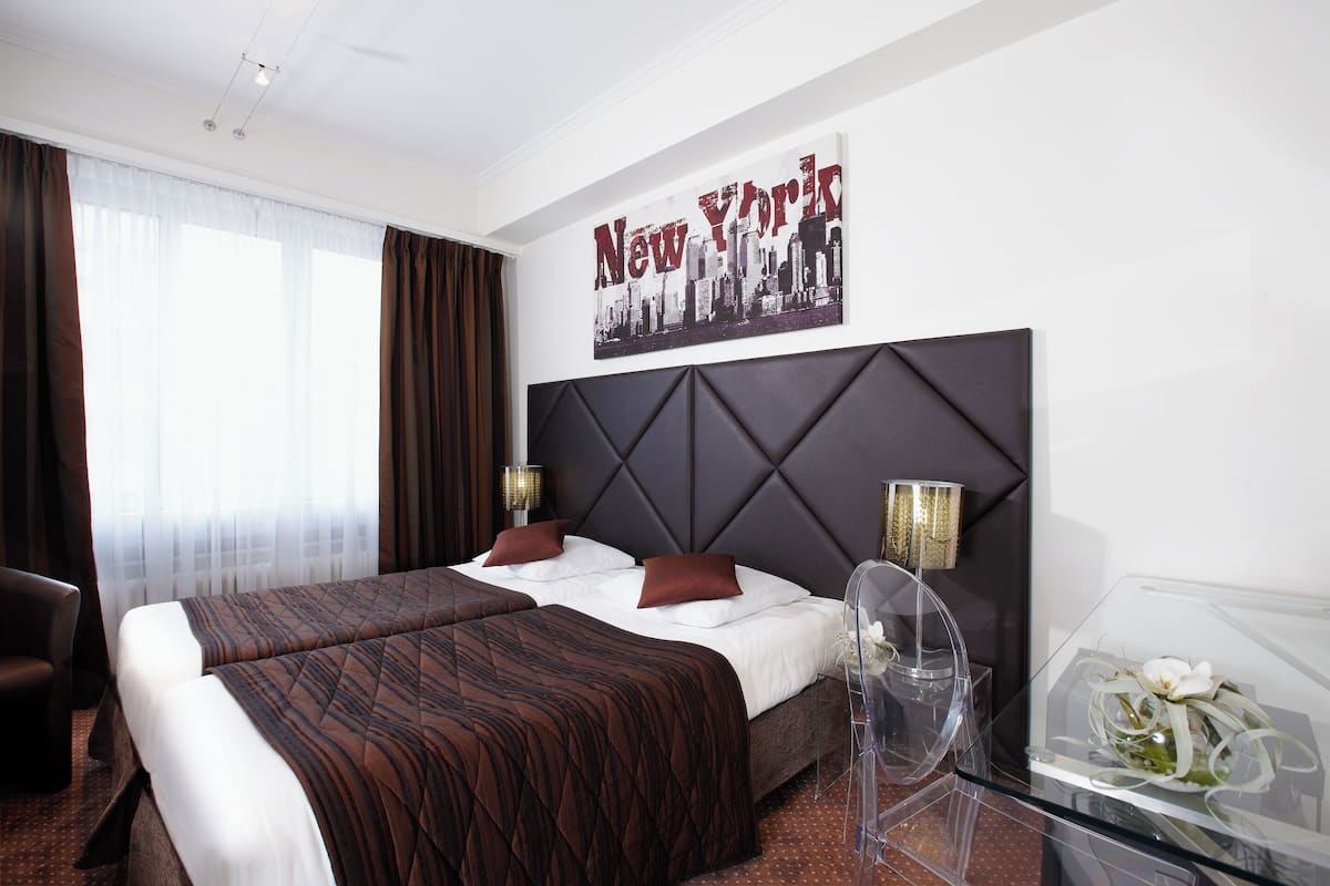 A hotel room with two beds, a cityscape poster, a modern headboard, a clear glass table, and a chair.