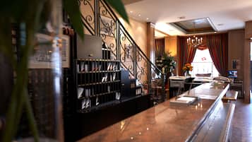 A hotel lobby with a marble reception desk, a staircase with ornate railings, and a large window with curtains.