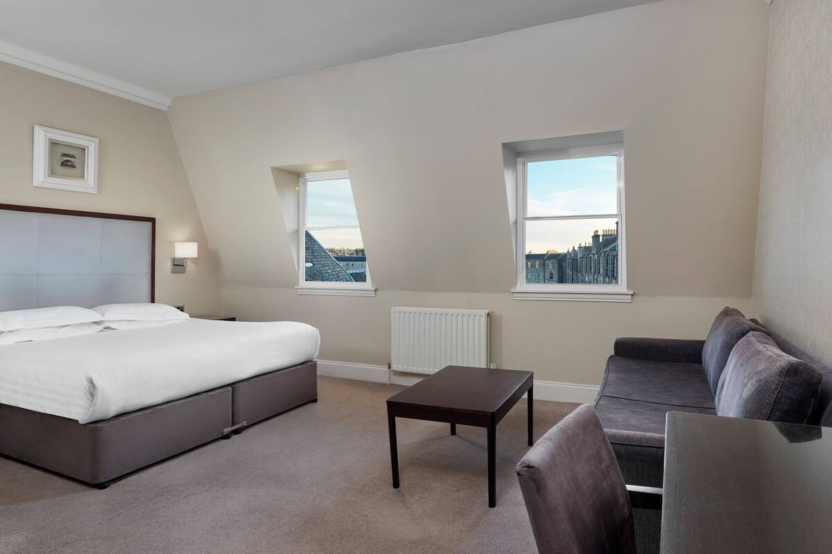 A bedroom with a bed, a sofa, a small table, and two windows offering city views.