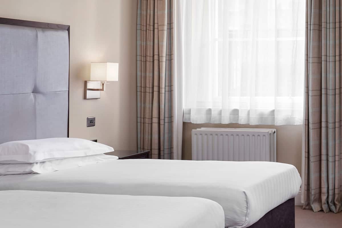 A hotel room with two beds, a wall-mounted light, a radiator, and plaid curtains.