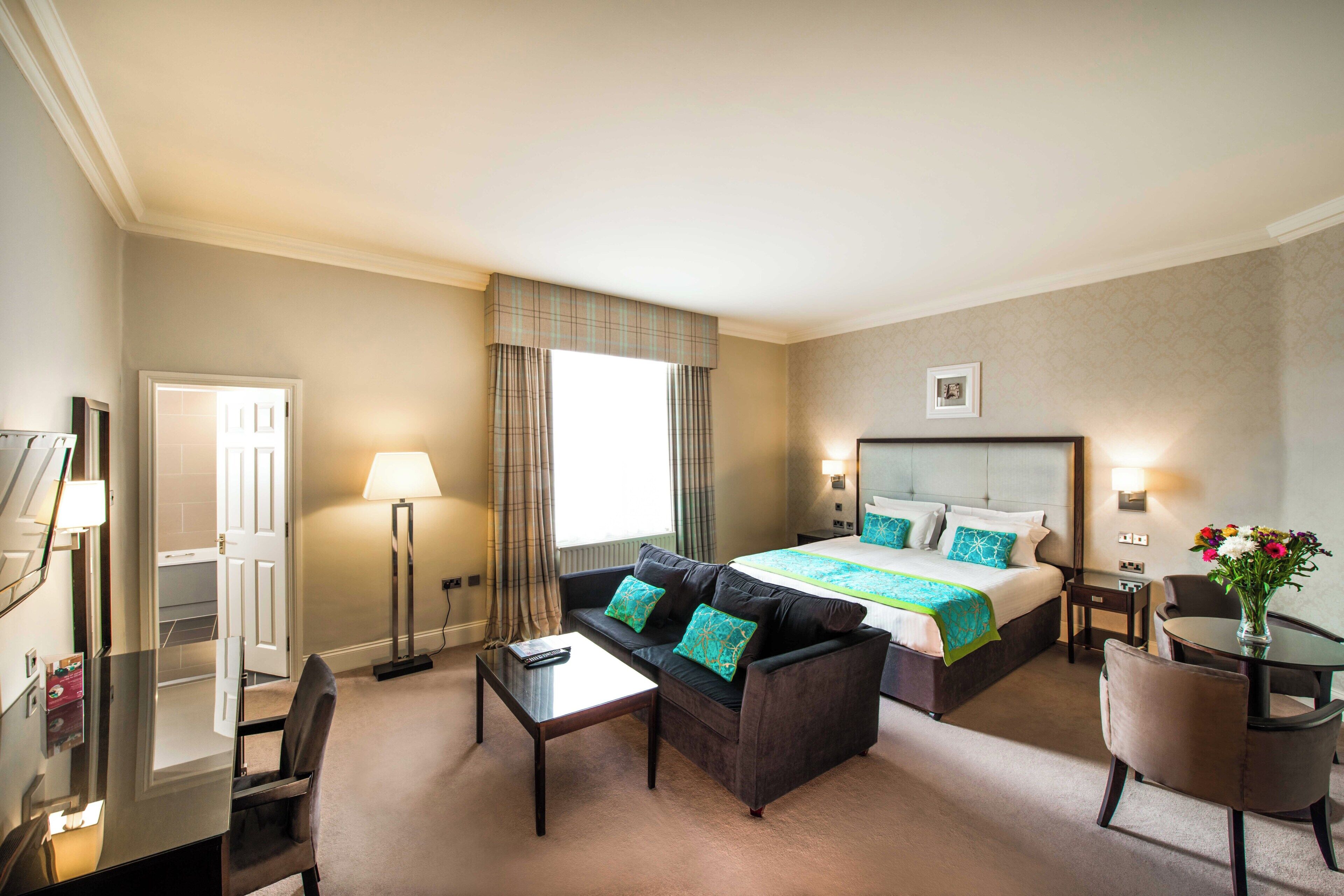 A hotel room with a bed, a sofa, a desk, a chair, a lamp, and a vase with flowers.