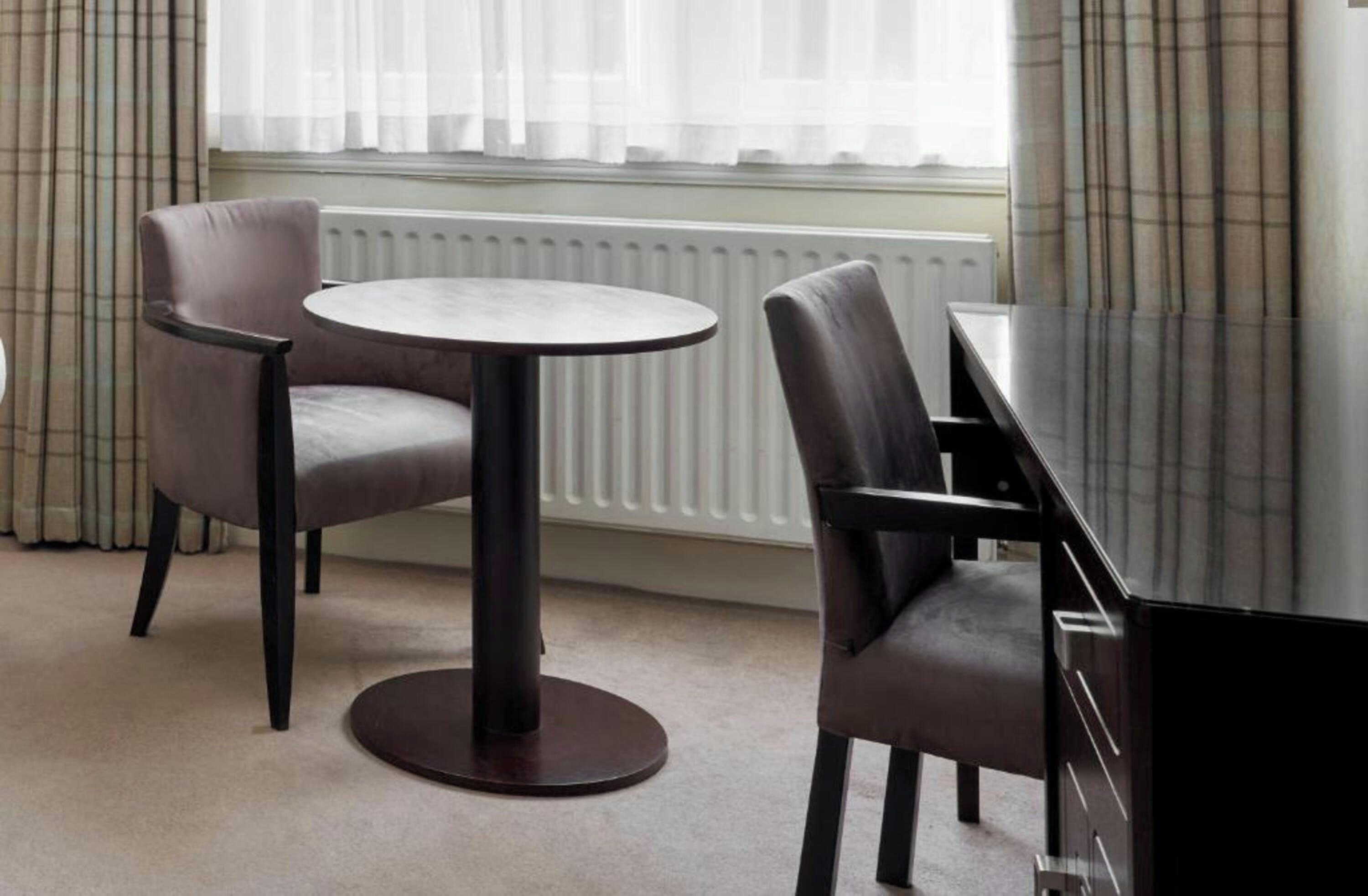 A hotel room with a round table and two chairs, a radiator, and a window with curtains.