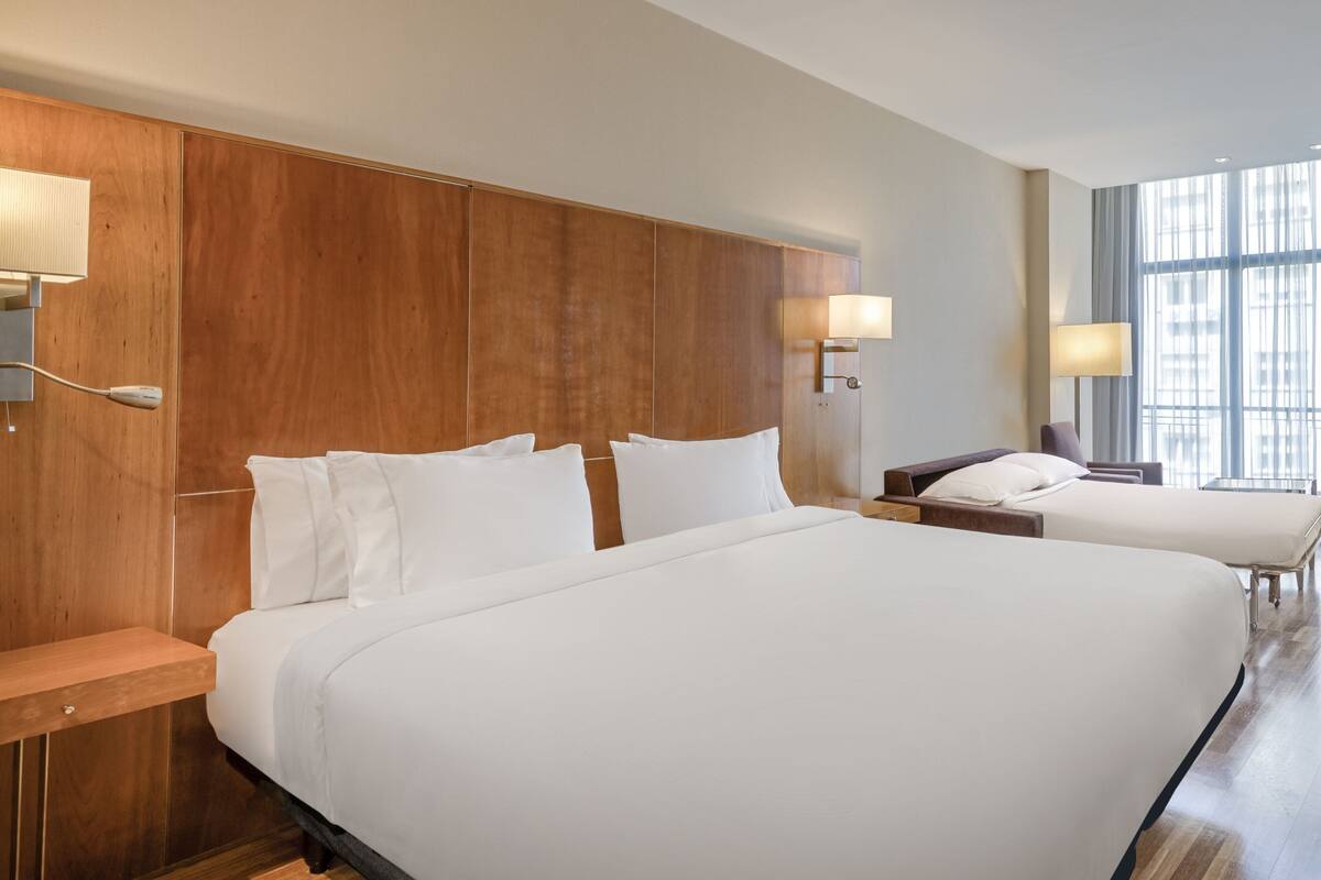 Une chambre d’hôtel avec deux lits, une tête de lit en bois, des tables de chevet, des lampes et une grande fenêtre.