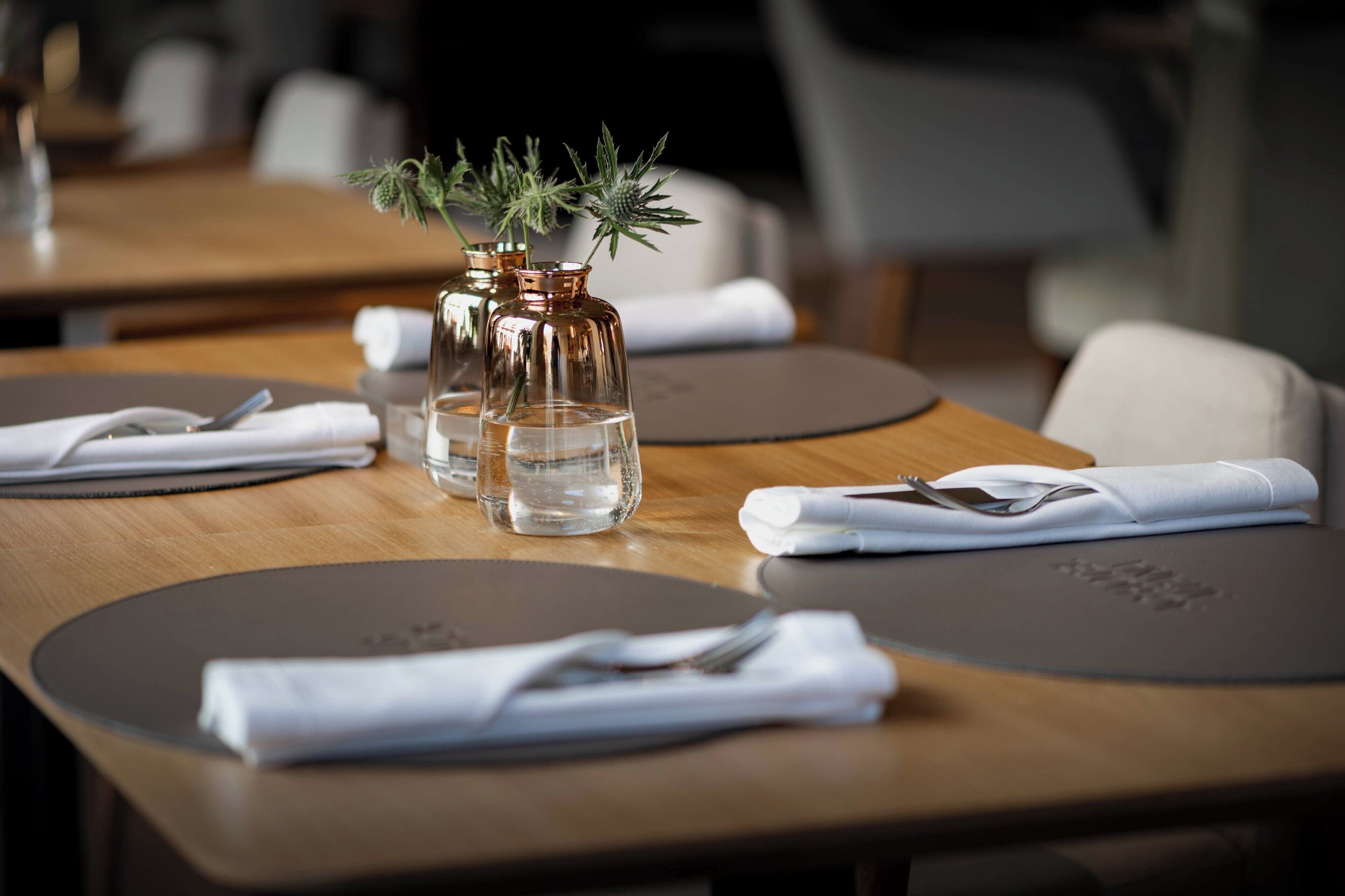 Una mesa con un mantel individual, una servilleta doblada, un tenedor, un cuchillo y dos jarrones de vidrio transparente con agua y plantas verdes.