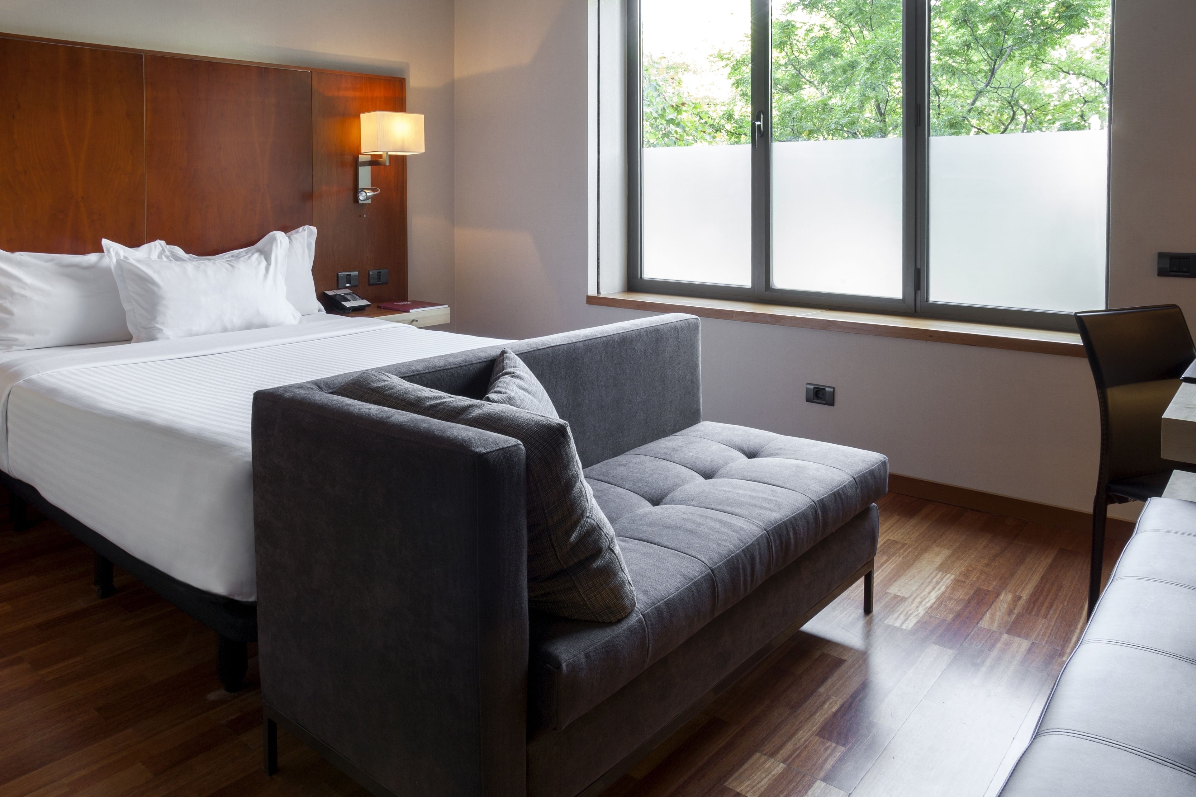 Une chambre d’hôtel avec un grand lit, un canapé gris, une tête de lit en bois, une table de chevet avec une lampe et une fenêtre donnant sur des arbres.