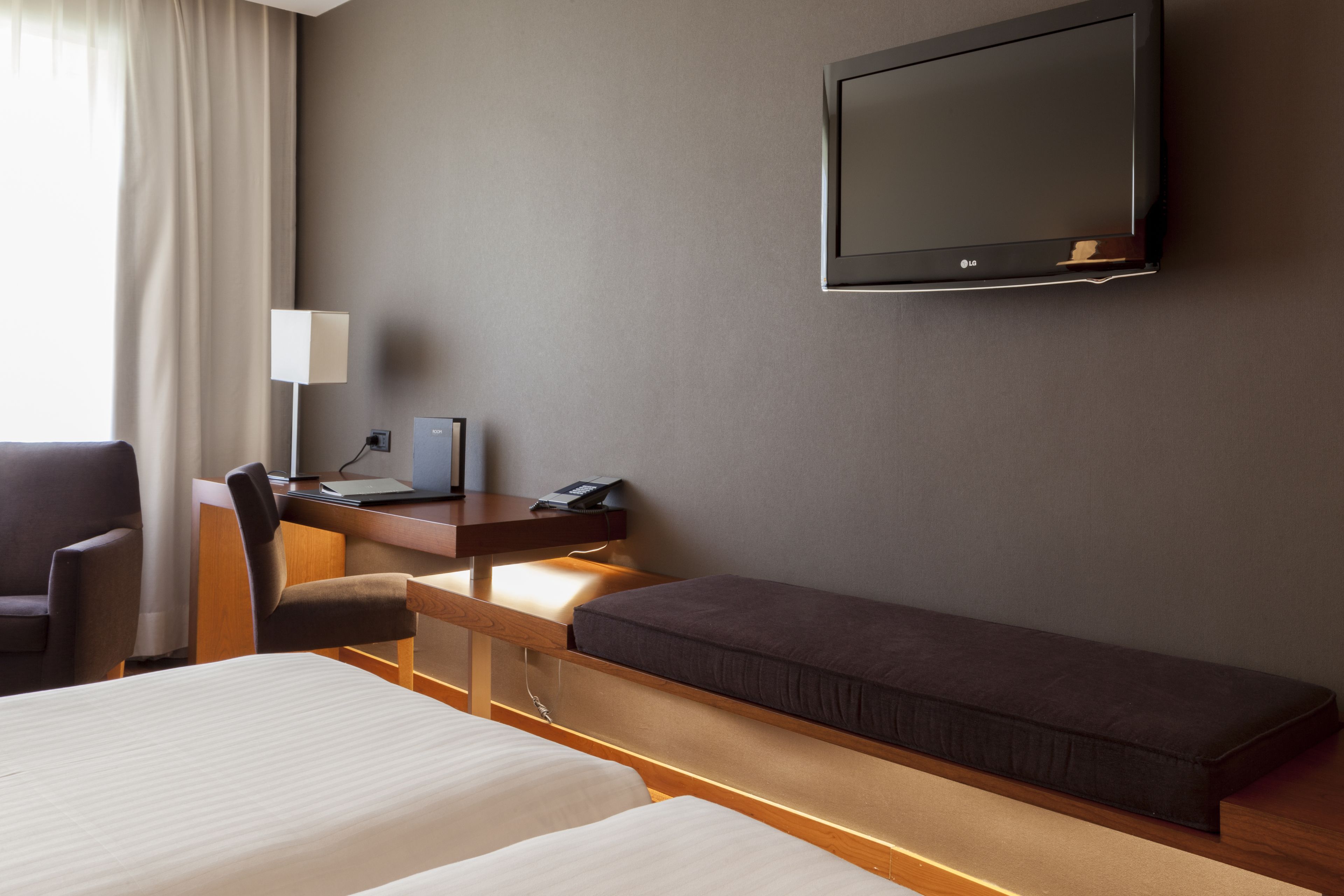 A hotel room with two beds, a desk with a computer, a chair, a TV mounted on the wall, and a lamp.