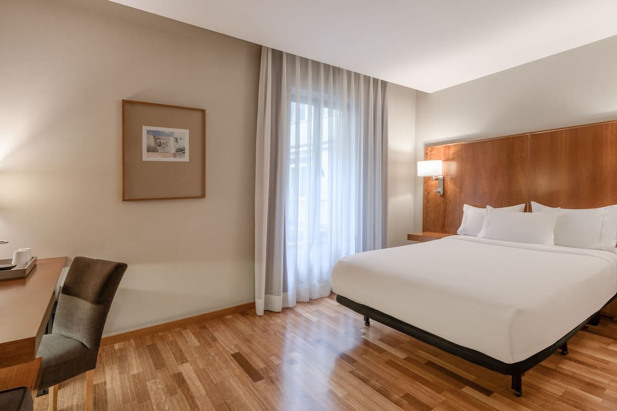 Une chambre d’hôtel avec un grand lit, une tête de lit en bois, un bureau avec une chaise, un tableau encadré au mur et des rideaux légers à la fenêtre.