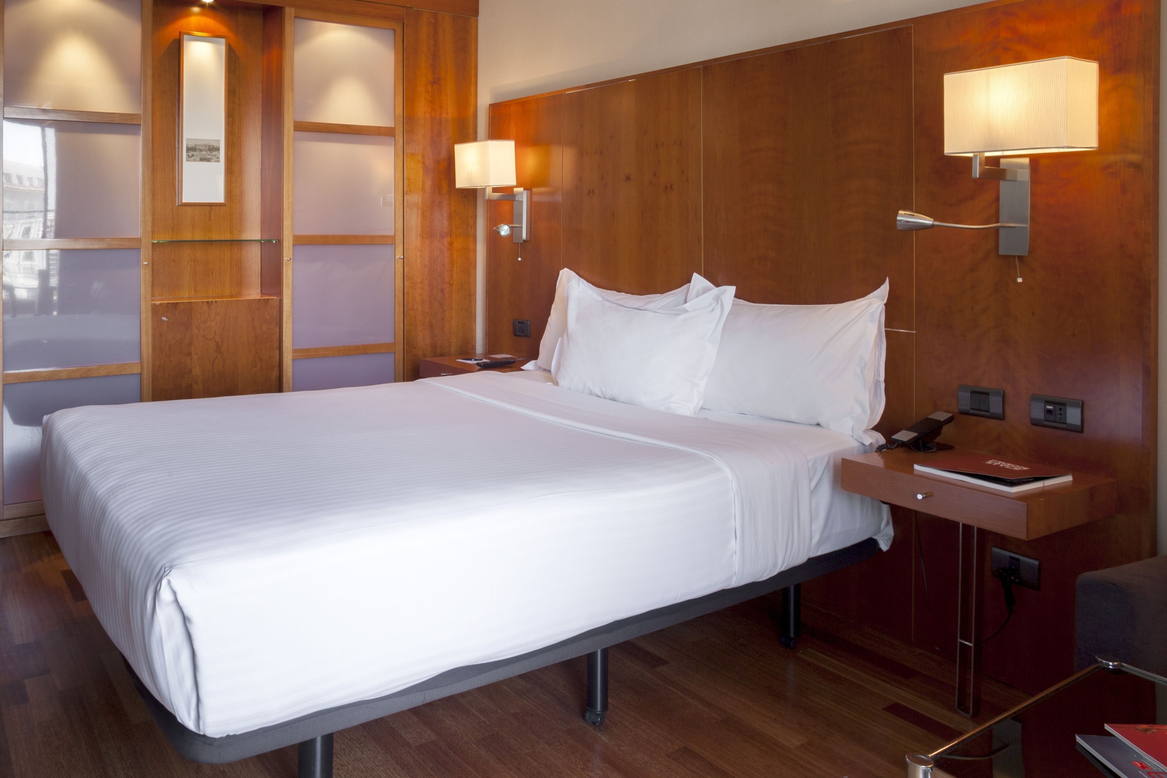 Ein Hotelzimmer mit einem großen Bett, einem Holz-Kopfteil, zwei Nachttischlampen, einem Nachttisch mit Telefon und einem kleinen Tisch mit einem Buch.