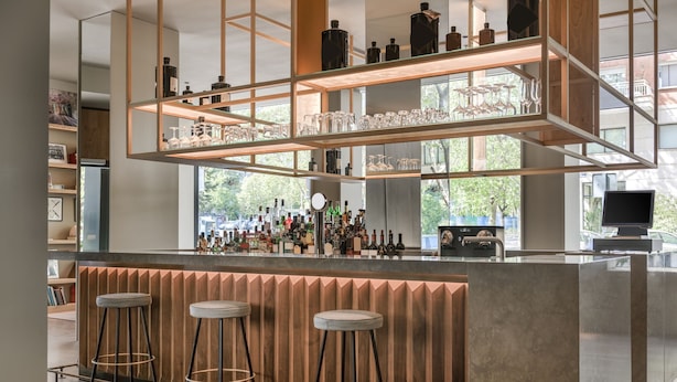 Un área de bar moderna con mostrador de madera, varios taburetes y estantes con botellas y copas.