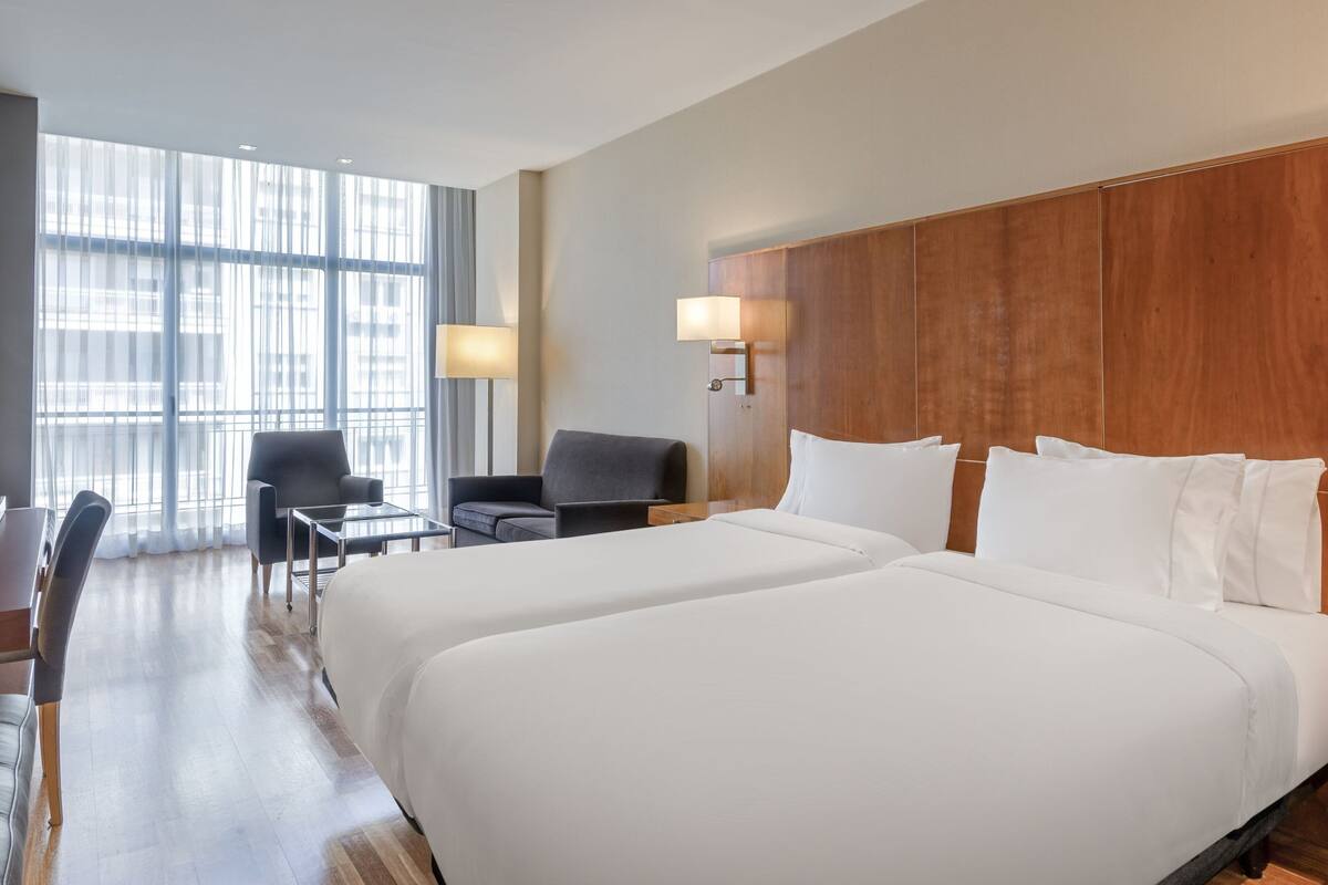 Une chambre d’hôtel avec deux grands lits, un bureau, une chaise et une grande fenêtre avec des rideaux.