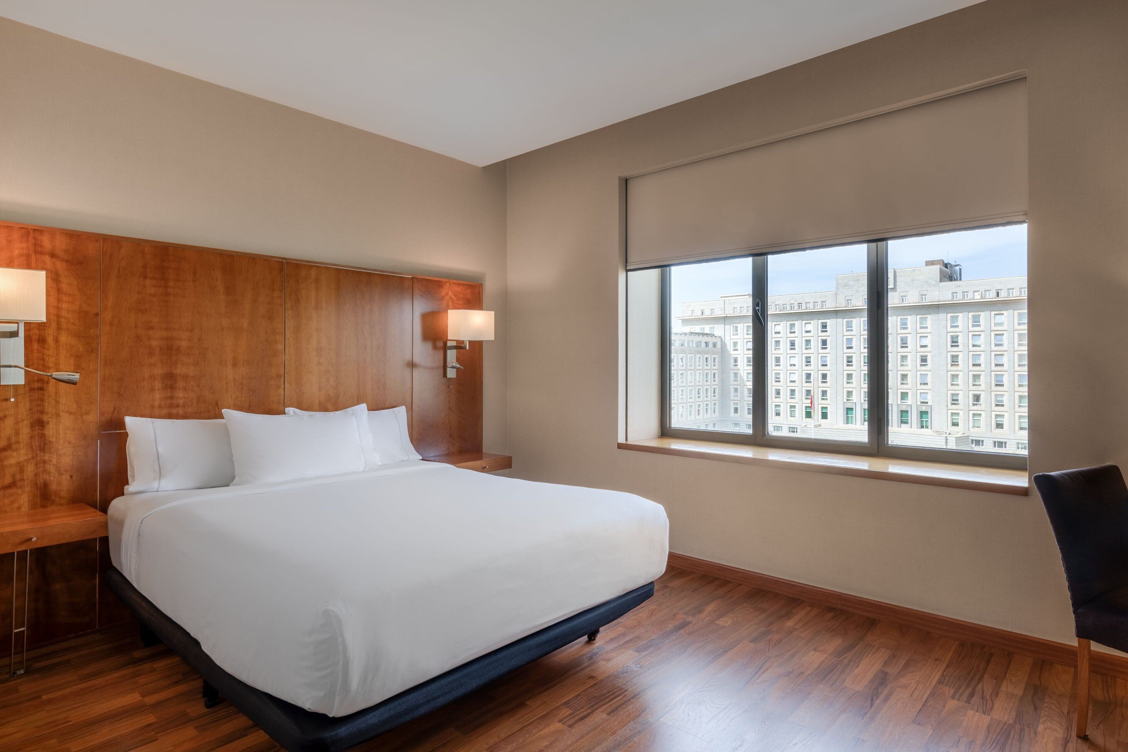 A hotel room with a large bed, wooden headboard, two bedside lamps, a window with a view of buildings, and a chair.