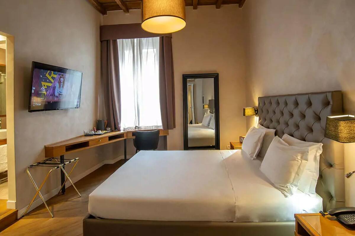 A hotel room with a bed, a desk, a chair, a television, a mirror, and a lamp.