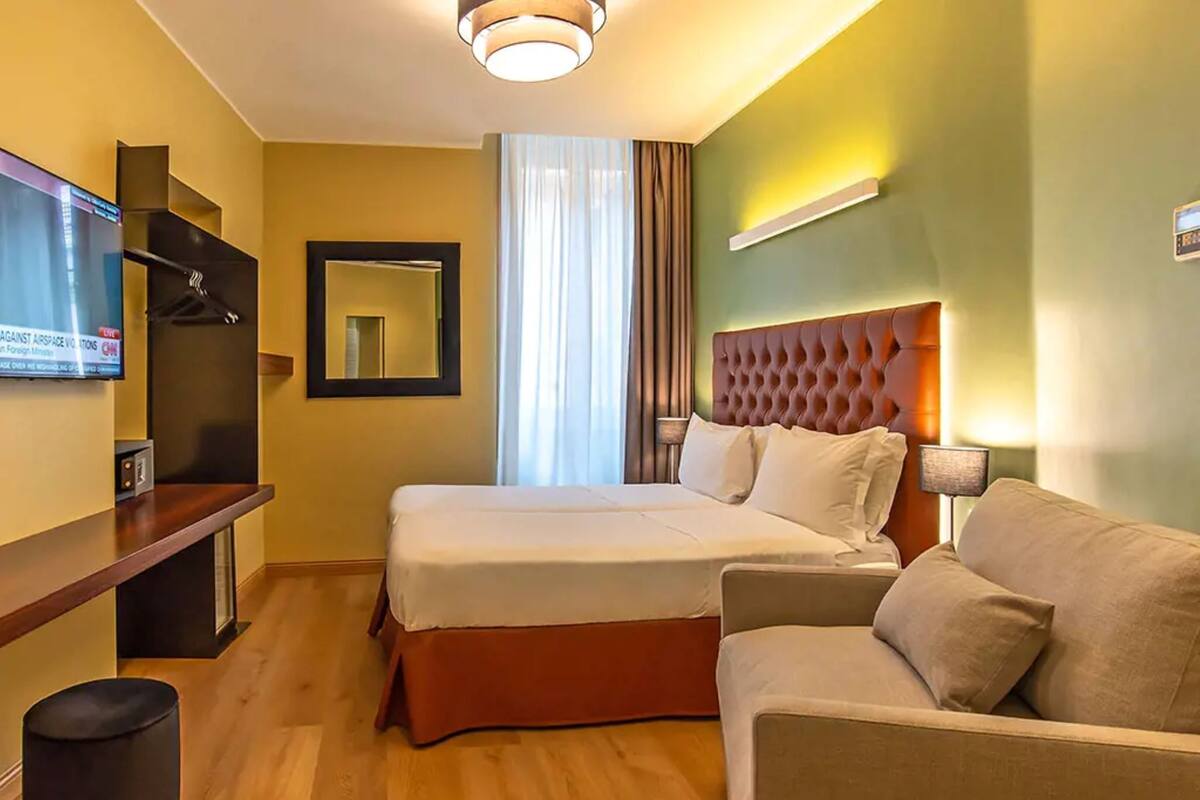 A hotel room with a bed, a sofa, a TV, a mirror, and a lamp.