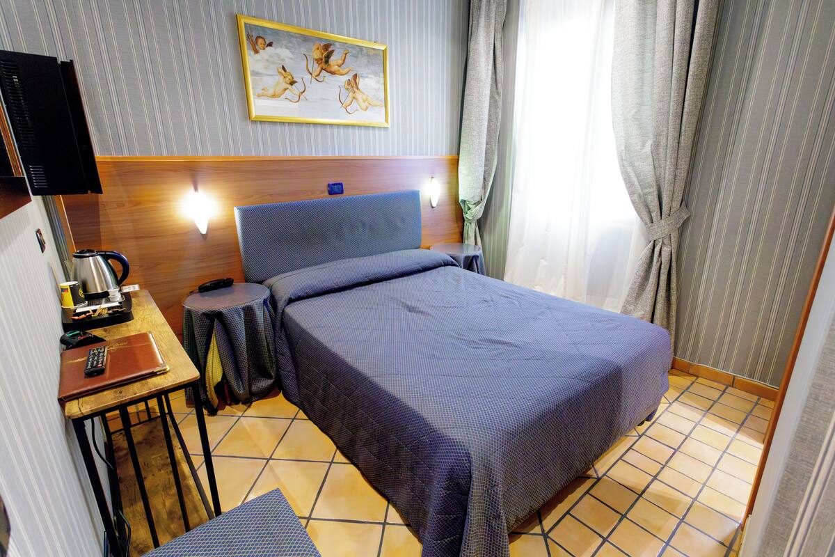 Una camera d'albergo con un letto, una scrivania con un portatile, una macchina del caffè e un quadro appeso al muro.
