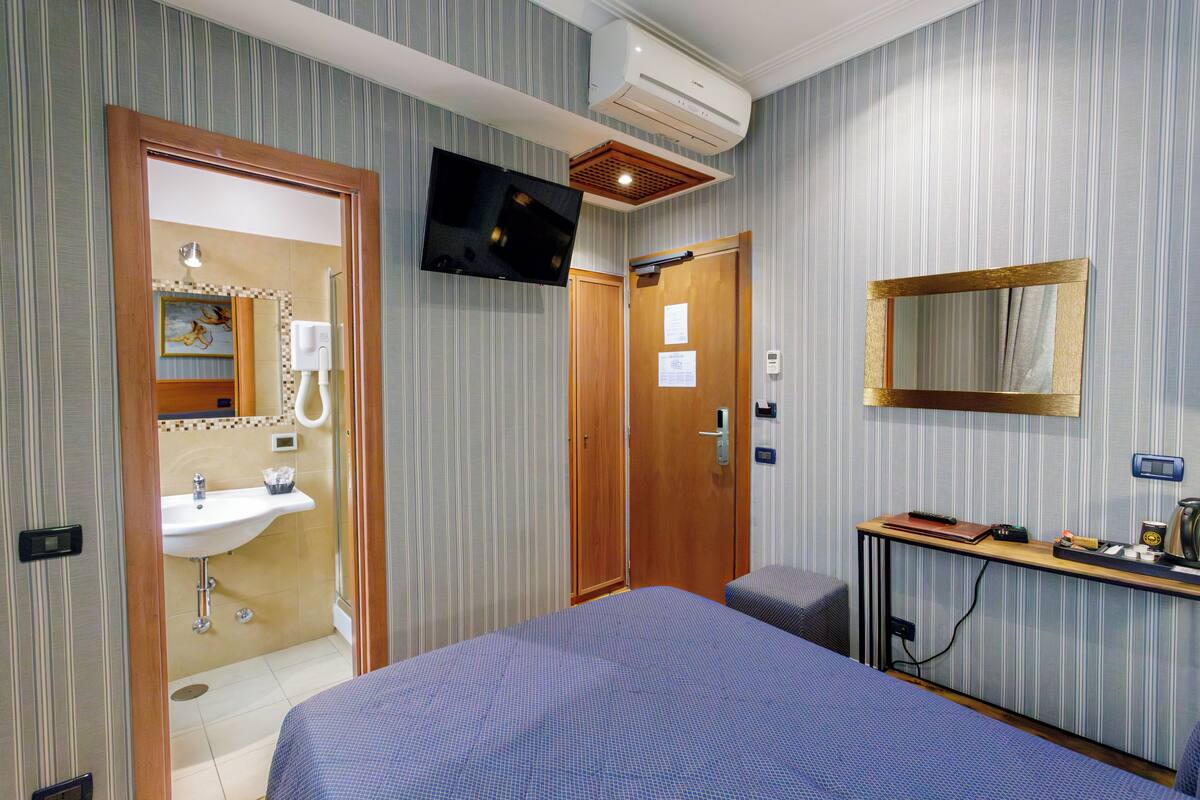 Una camera d'albergo con un letto, un comodino con una lampada, una TV a parete e un ingresso che conduce al bagno.