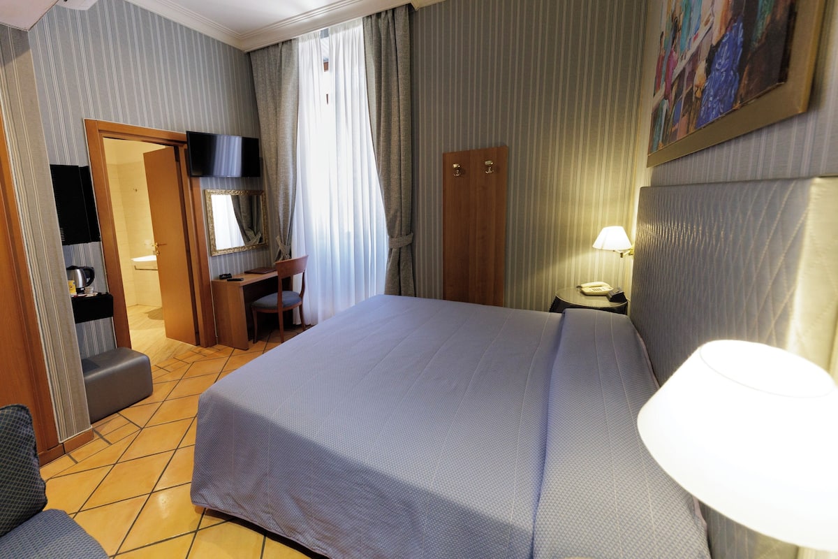 Una camera d'albergo con un letto grande, una scrivania, una sedia, un televisore, una lampada e un bagno.
