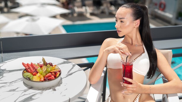 A person sitting at a table with a fruit bowl and a drink, overlooking a pool area with umbrellas and lounge chairs. - Capsis Hotel Thessaloniki (Thessaloniki)